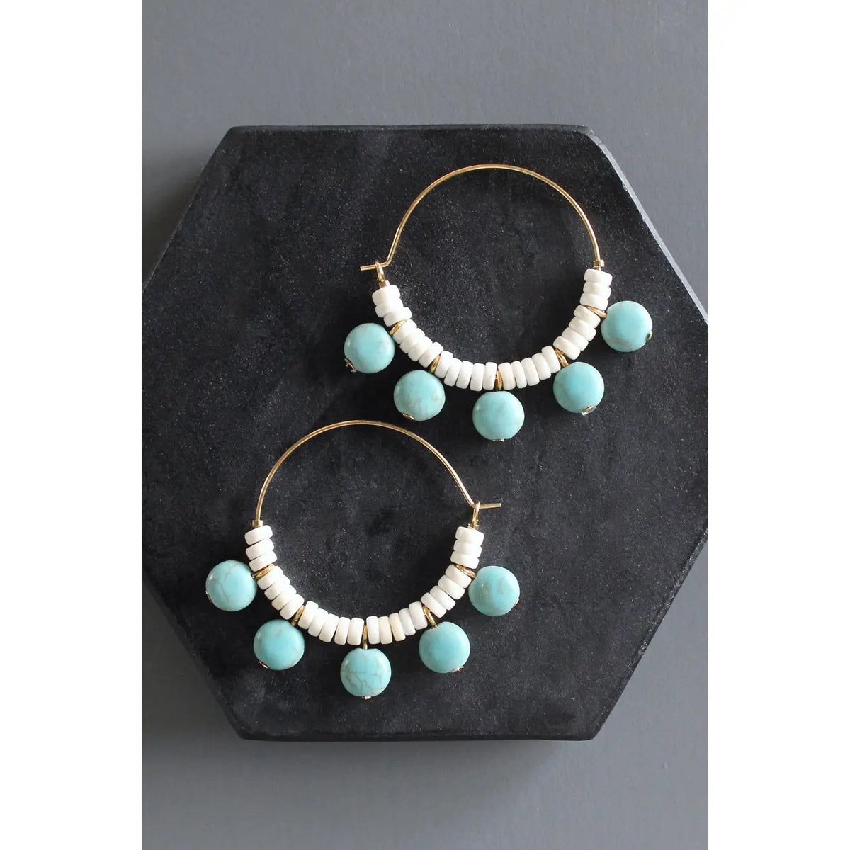 DAVID AUBREY - TUQUOISE AND WHITE HOOP EARRINGS by DAVID AUBREY, featuring gold plated hoops embellished with alternating white magnesite heishi beads and round turquoise glass beads, are displayed on a black hexagonal surface against a dark gray background.