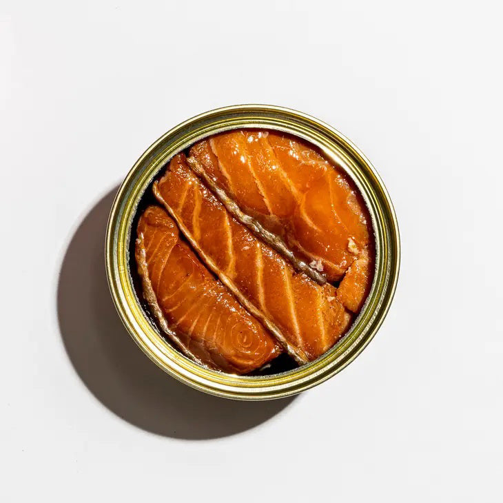 A vibrant red tin of FISHWIFE - SMOKED ATLANTIC SALMON features a colorful woman with fish and lemons, proudly stating "100% Traceable," "Responsibly Raised in Norway," and honoring sustainable seafood, from the brand FISHWIFE.