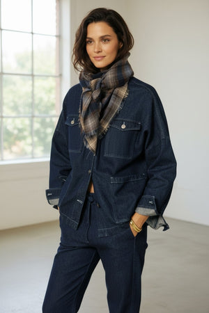 A woman stands indoors near a large window with natural light, wearing a dark denim jacket and pants, accessorized with the HARTFORD checked wool scarf in navy by HARTFORD.