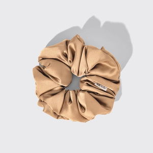 A package of KITSCH PILLOW SATIN SCRUNCHIES IN BLUSH sits behind four stacked scrunchies in beige, pink, cream, and champagne shades. The packaging displays a woman’s face and reads “Pillow Scrunchies in Satin” by KITSCH, showcasing this gentle, damage-free hair accessory ideal for soft styling.