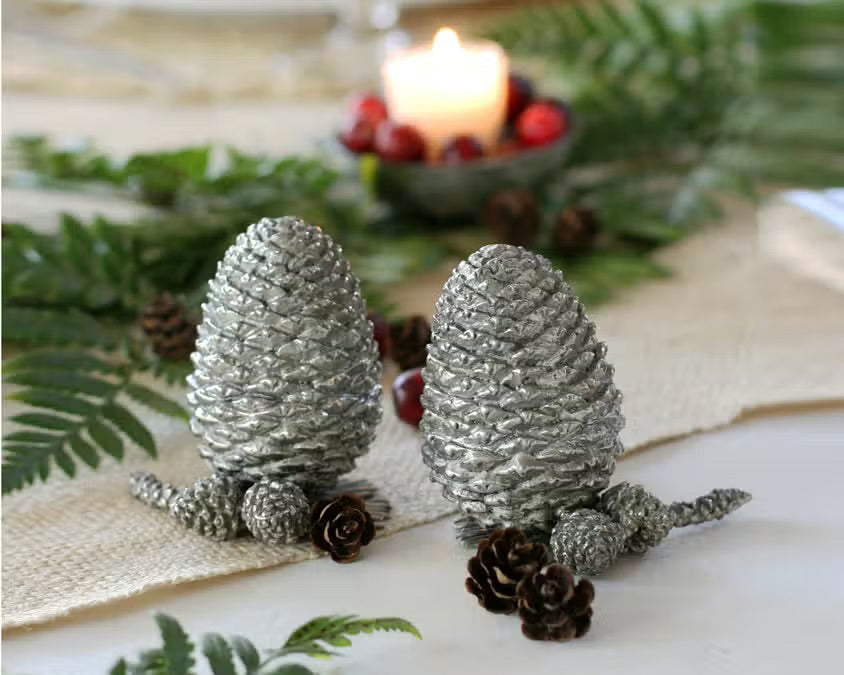 Two detailed silver pine cones stand upright on a white background, resembling the VAGABOND HOUSE - PEWTER PINE CONESALT AND PEPPER SET by VAGABOND HOUSE, with small pine needle segments visible at their bases.