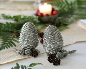 Two detailed silver pine cones stand upright on a white background, resembling the VAGABOND HOUSE - PEWTER PINE CONESALT AND PEPPER SET by VAGABOND HOUSE, with small pine needle segments visible at their bases.