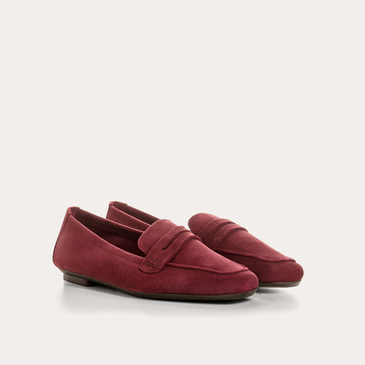 Close-up of the REQINS - HEMA SUEDE LOAFER IN BORDEAUX by REQINS, showcasing artisanal craftsmanship with burgundy suede, detailed stitching, a front strap, and a soft leather insole, displayed on a light surface.