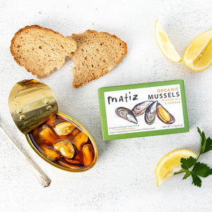 A green rectangular box labeled "MATIZ - MUSSELS IN ORGANIC OLIVE OIL" from MATIZ ESPANA. The box features illustrations of three mussels, one of which is open to show the yellow inside. It also notes "0.8 grams of Omega-3s per serving" and weighs 111g (4 oz).