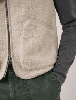 A man is wearing the HARTFORD - BASTA SHERPA VEST IN NATURAL over a gray long-sleeve shirt, holding the vest’s zipper with one hand. The plain, light-colored background complements the cozy, Hartford-inspired style of this men’s vest from HARTFORD.