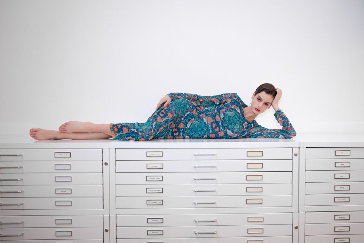 The FRANCO FERRARI BALLET MESH DRESS is a long-sleeved, fitted dress with a green, teal, and orange botanical print, semi-sheer fabric, a round neckline, and an intricate symmetrical floral pattern.