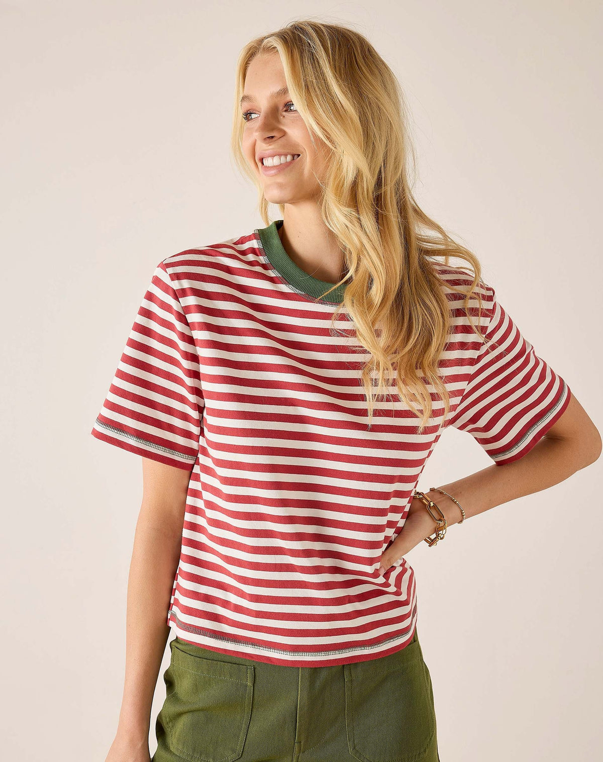 A smiling person with long blonde hair wears the MERSEA Deauville Ringer Tee in Currant Jade—a red and white striped ringer tee with green collar and cuffs—paired with green pants, standing against a light background.