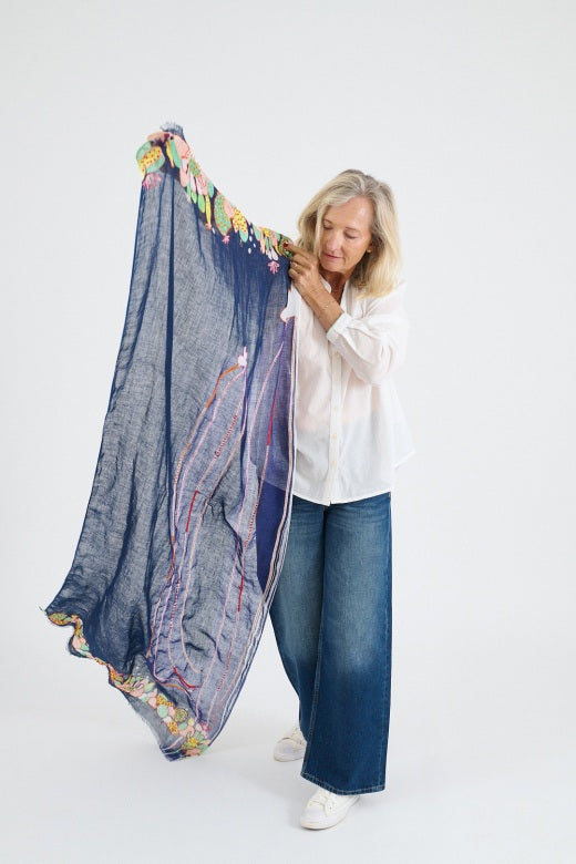 A blonde woman in a white blouse and blue jeans stands against a plain background, wearing the STORIATIPIC AZUR SCARF IN MARINE—100% linen with colorful handmade cactus embroidery.