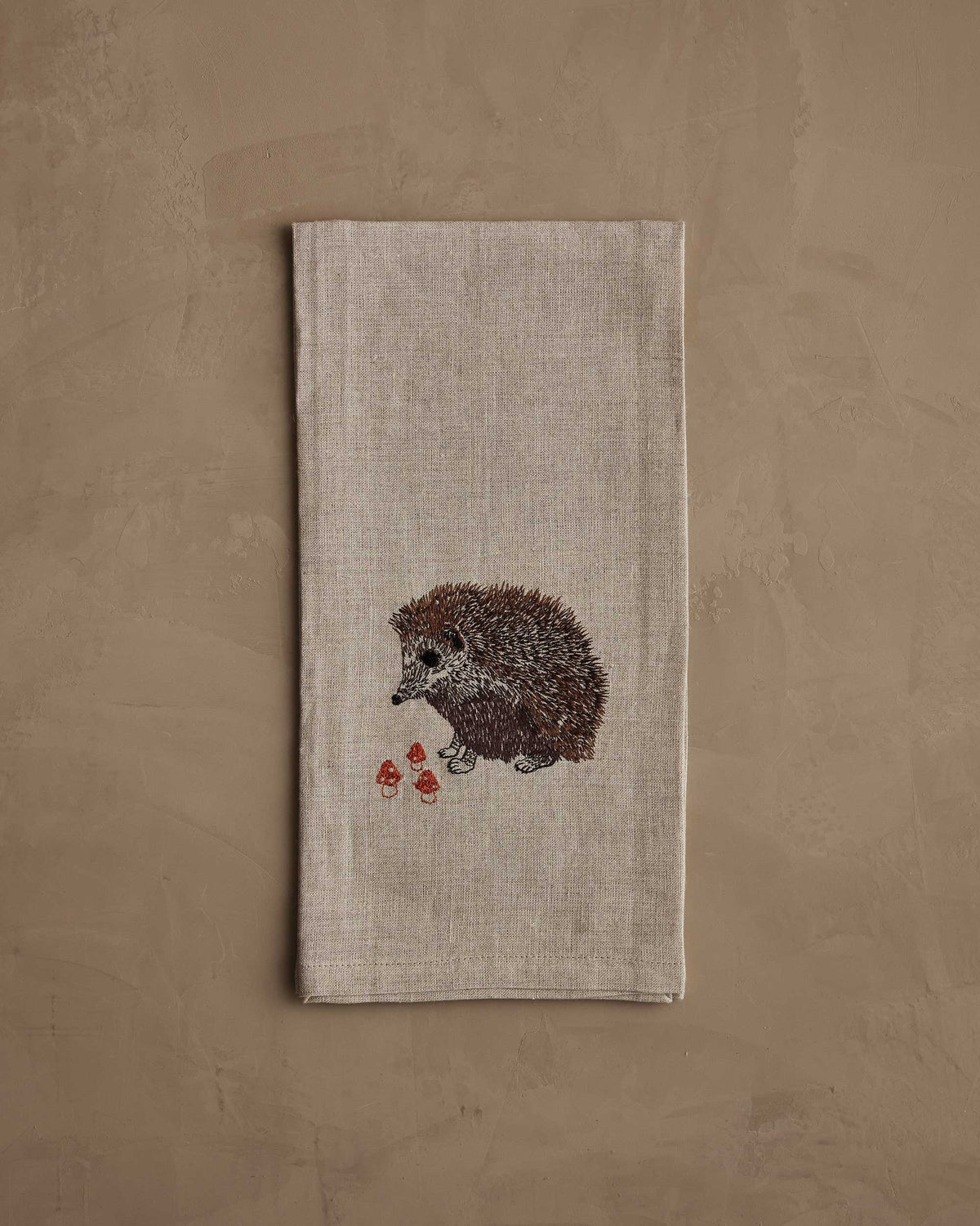 A HEDGEHOG WITH MUSHROOMS LINEN TEA TOWEL by CORAL & TUSK hangs from the handle of a white stove in a kitchen.