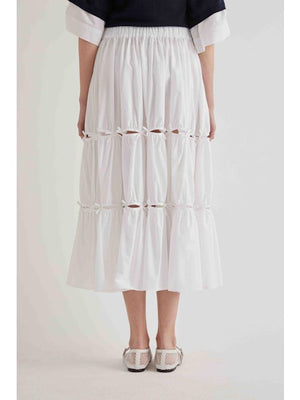 A person stands sideways in a MEIMEIJ cotton poplin midi skirt with bow detail and gathered tiers, paired with a black rolled-sleeve top and white mesh ankle-strap shoes. The background is plain and light-colored.