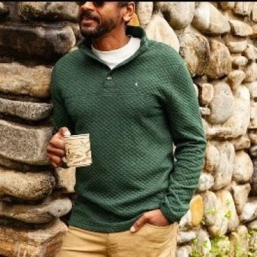 Wearing the CRIQUET Quilted Pullover in Heather Ivy, a person stands by a stone wall in khaki pants, holding a patterned mug and smiling, with greenery in the background—highlighting this classic-fit all-season layer from CRIQUET.