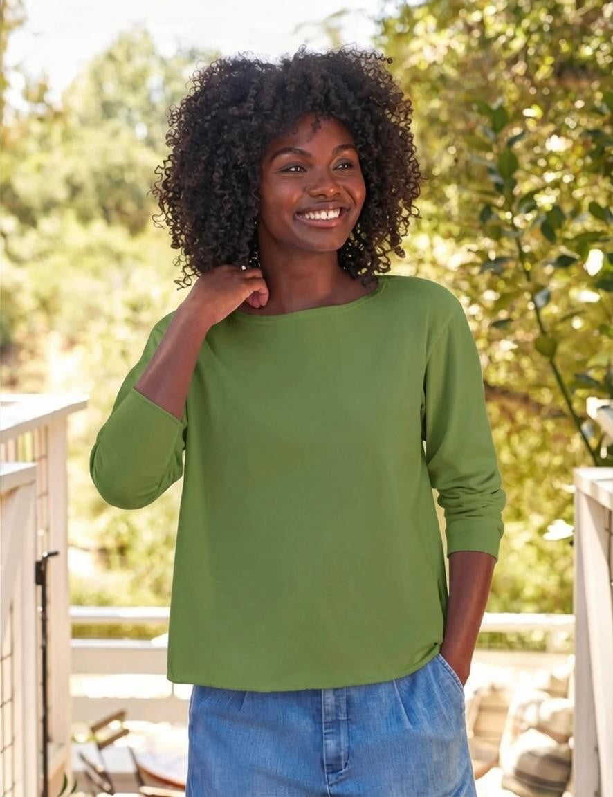 A woman with curly hair smiles outdoors on a sunlit deck surrounded by greenery and trees, wearing the FRANK & EILEEN ELLA Long Sleeve Boatneck Tee in Nori—proudly made in California—and blue denim pants.