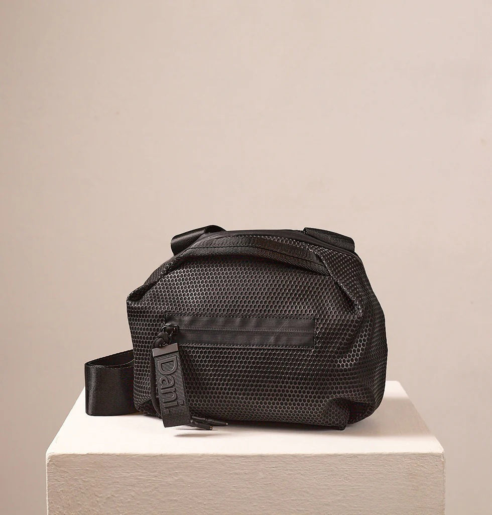 A person in a light beige suit carries the DANIELLA LEHAVI - BONNIE TRAVEL CROSSBODY, a large brown vegan bag with black zipper and strap, shown from shoulders to mid-thigh against a plain background.