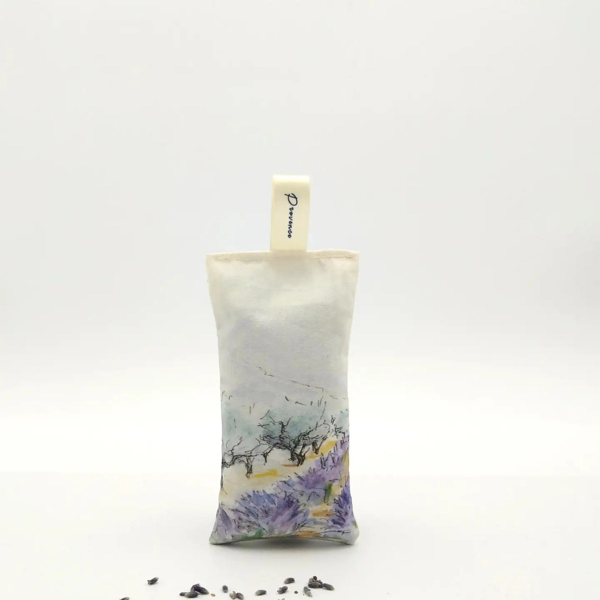 A rectangular L’AUGUSTE PROVENCE Lavender Sachet from Provence, featuring a scenic design and labeled fabric loop, stands upright on a white background with scattered Provence lavender buds in front.