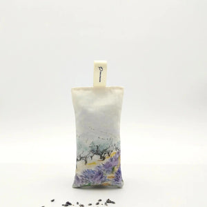 A rectangular L’AUGUSTE PROVENCE Lavender Sachet from Provence, featuring a scenic design and labeled fabric loop, stands upright on a white background with scattered Provence lavender buds in front.