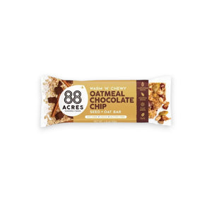 A packaged snack bar labeled "88 ACRES - OATMEAL CHOCOLATE CHIP SEED OAT BAR" from the brand 88 ACRES is shown against a light wooden background. The packaging highlights that it is a seed + oat bar, emphasizing its nut-free, vegan, and gluten-free qualities, and promises creamy chocolate chips for a delightful chewy texture.