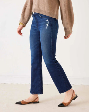 A person stands with hands in pockets against a plain light background, wearing MERSEA’s Murphy Mini Boot Jeans in Patriot—a pair of mid-rise, blue flare jeans crafted from four-way stretch denim—paired with a tan long-sleeve top, black pointed-toe flats, and gold bangles.