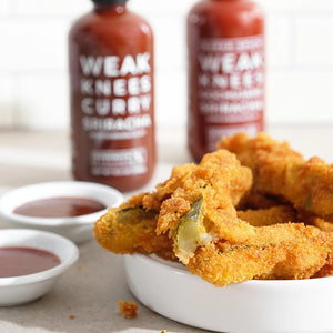 A 10.5 oz (298g) bottle of Bushwick Kitchen's 'Weak Knees' Curry Sriracha sauce, featuring a black cap and a white label prominently displaying "WEAK KNEES CURRY SRIRACHA BORN IN BROOKLYN, NY" in large bold letters, offers a unique twist on classic sriracha with its curry flavor.