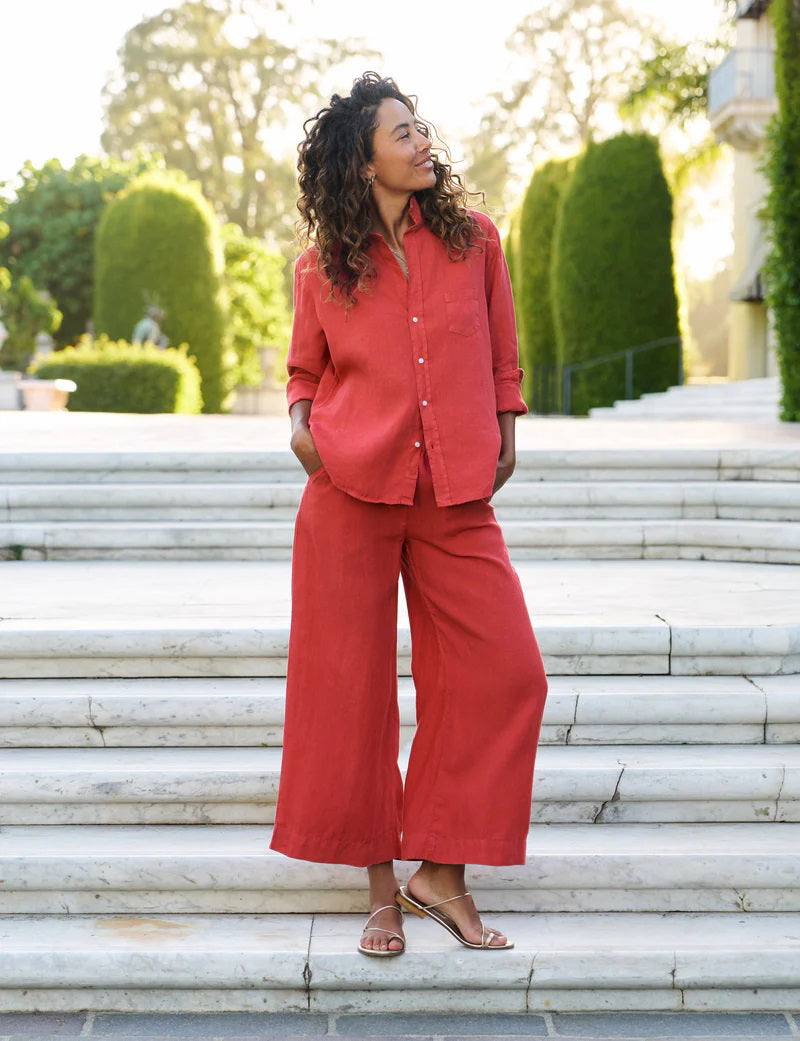 On marble steps outdoors, a woman with curly hair wears gold sandals and the FRANK & EILEEN Maisie Wide Leg Pull On Pant from the Tuscany Getaway Set in Summer Red. She stands with hands in her pockets, framed by green trees and manicured bushes.