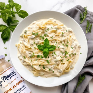 A bowl of green beans topped with creamy YO MAMA'S - ROASTED GARLIC ALFREDO sauce and crispy onions sits on a gray-striped napkin, beside a jar of YO MAMA'S FOODS Garlic Alfredo sauce and white salad servers.