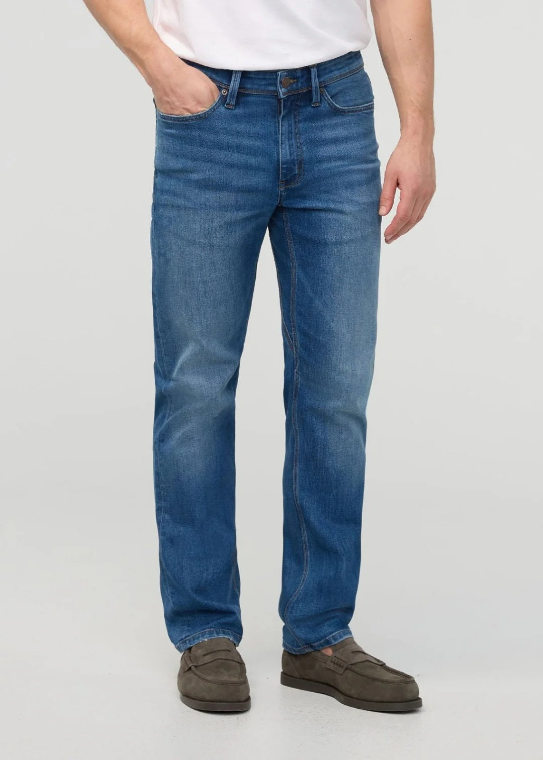 Close-up of a person wearing DUER’s PERFORMANCE DENIM+ Straight Pants in Alpine Indigo, showing relaxed hands and highlighting the texture, stitching, and stretch comfort of these blue jeans against a plain light background.