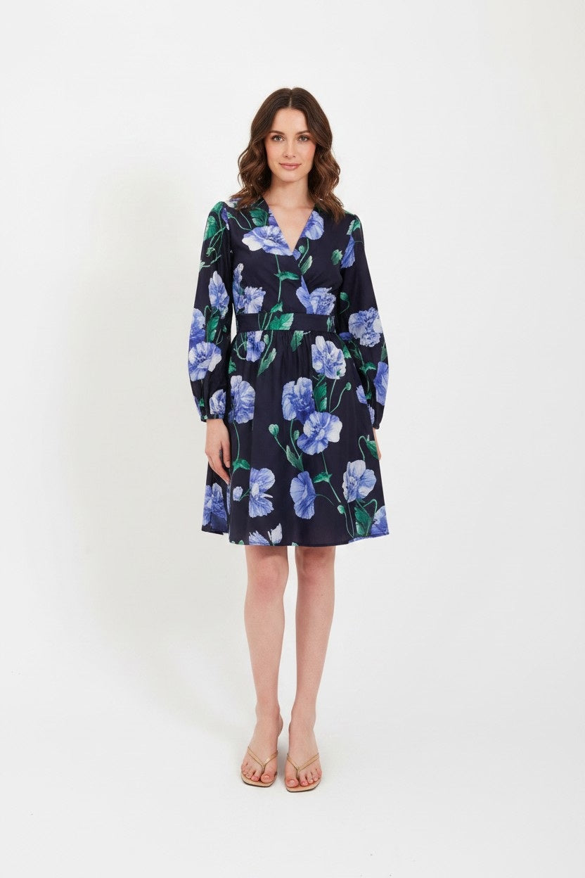 A woman wearing the LAVI Cetara Dress in Blue Poppies by LAVI—an Italian cotton, navy blue, long-sleeve dress featuring a green and blue botanical print—paired with beige sandals, stands against a plain white background.