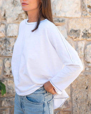 A woman with wavy brown hair models the MERSEA - CATALINA SLUB TEE—an oversized, long-sleeved white top with thin gray stripes—by MERSEA. She pairs it with blue jeans and stands against a plain, light background, hands behind her back.