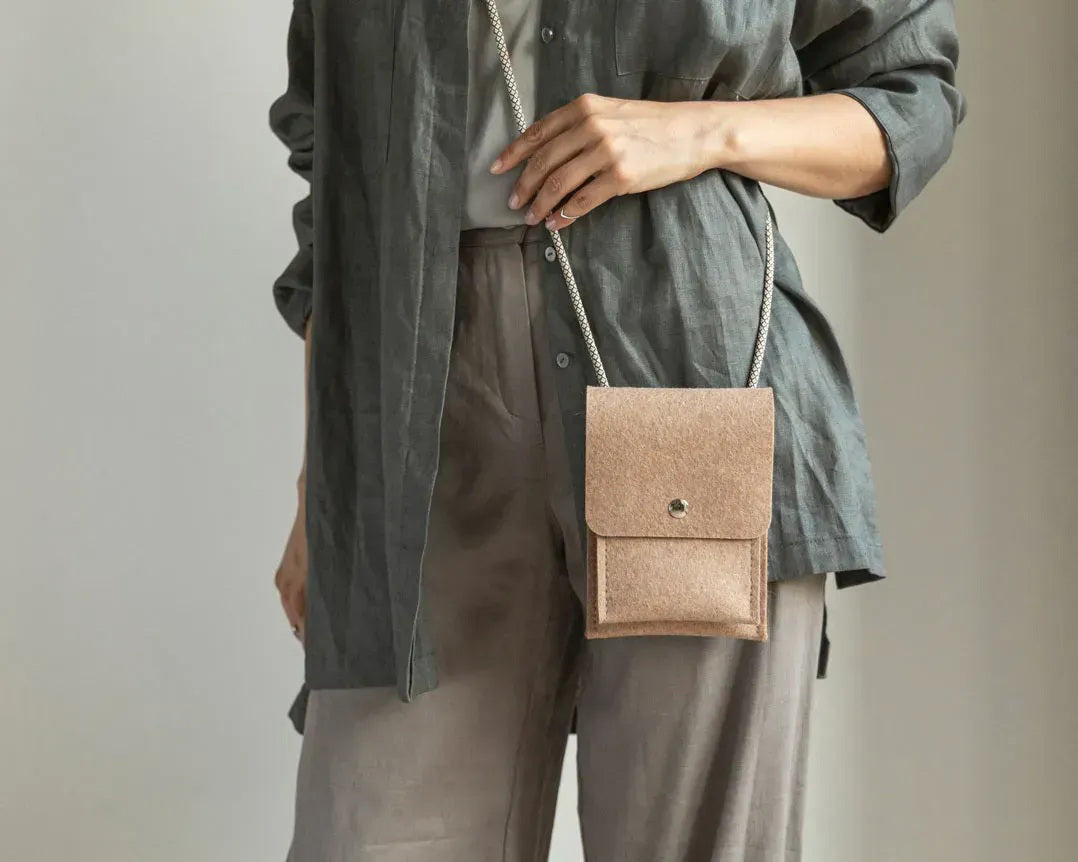 A person in a beige sweater and tan pants holds the WOOLBERRY WOOL FELT PHONE BAG, a small gray handcrafted bag with a flap and button closure. The image is cropped at the shoulders and knees.
