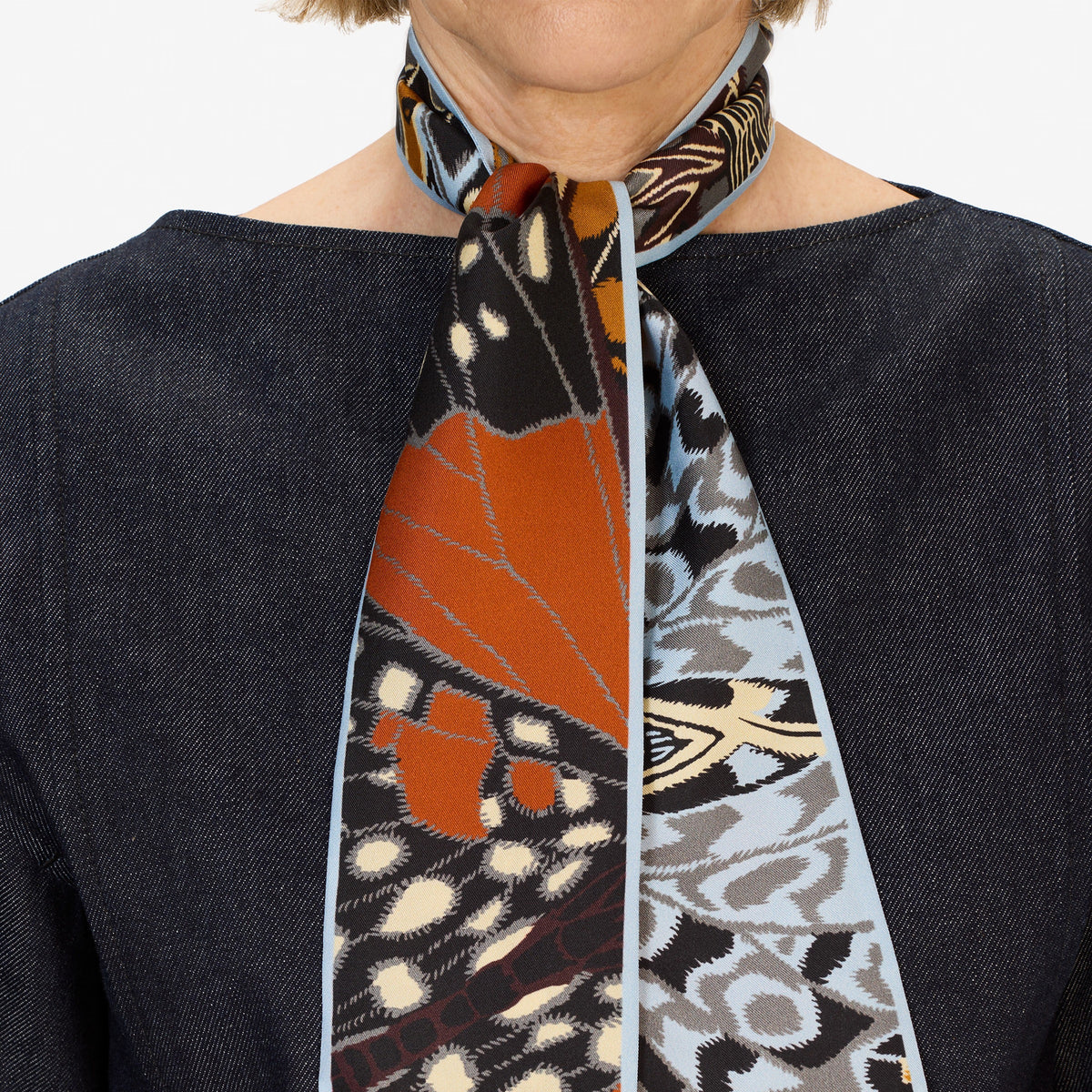 A woman wears a dark denim top paired with the INOUI EDITIONS - PAPILLON (BUTTERFLIES) silk ribbon by INOUI, styled as a chic neck accessory featuring butterfly and animal print patterns.
