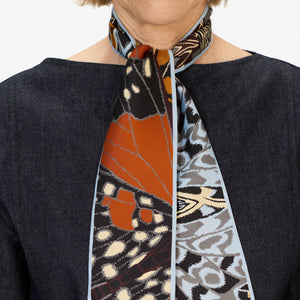 A woman wears a dark denim top paired with the INOUI EDITIONS - PAPILLON (BUTTERFLIES) silk ribbon by INOUI, styled as a chic neck accessory featuring butterfly and animal print patterns.