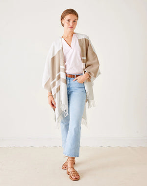 A person stands against a plain white background, wearing a light pink blouse and the MER SEA - CLASSIC TRAVEL WRAP IN DUNE STRIPE. Their light blue jeans and brown sandals finish the look. With their hair tied back, they strike a relaxed pose with one hand on their hip.
