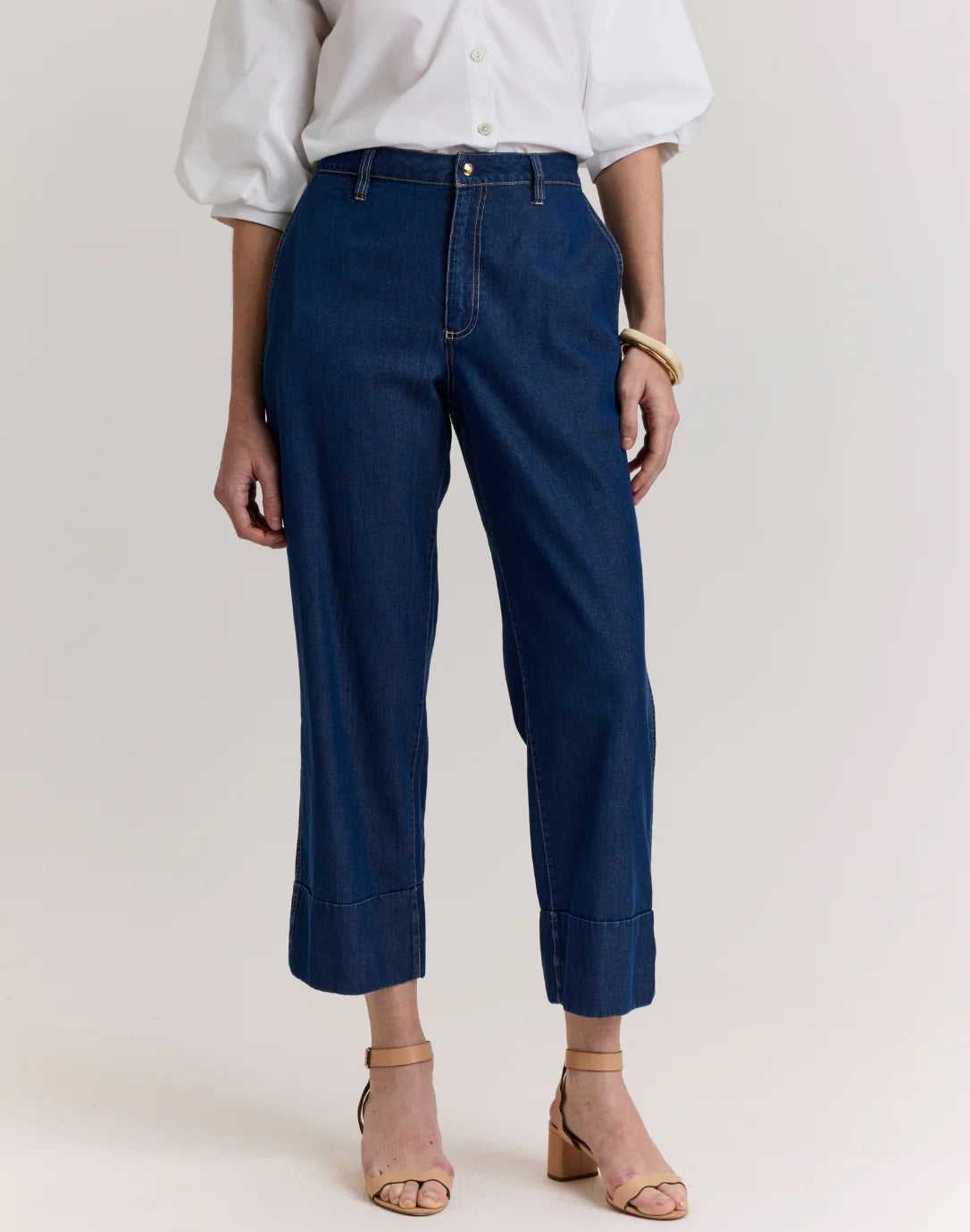 A person stands against a neutral background wearing a white button-up blouse, beige heeled sandals, and the HINSON WU - ROSA DARK WASH DENIM CROPPED PANT by HINSON WU.
