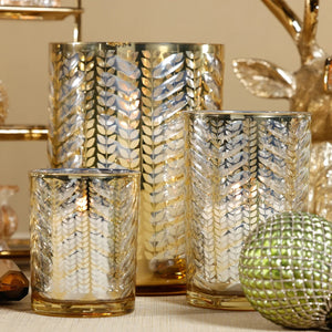 Three 4 x 5 inch LASER ETCHED METALIC HURRICANE 5″ cylindrical glass candle holders with a gold leaf pattern, each containing a lit candle.
