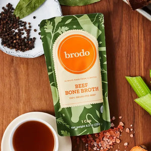 A pouch of BRODO BROTH- GRASS FEED BONE BROTH from BRODO BROTH COMPANY rests on a wooden surface, accompanied by a cup of collagen-rich broth, celery sticks, salt crystals, black peppercorns, and a bay leaf—ideal for sipping or crafting a rich pan sauce.