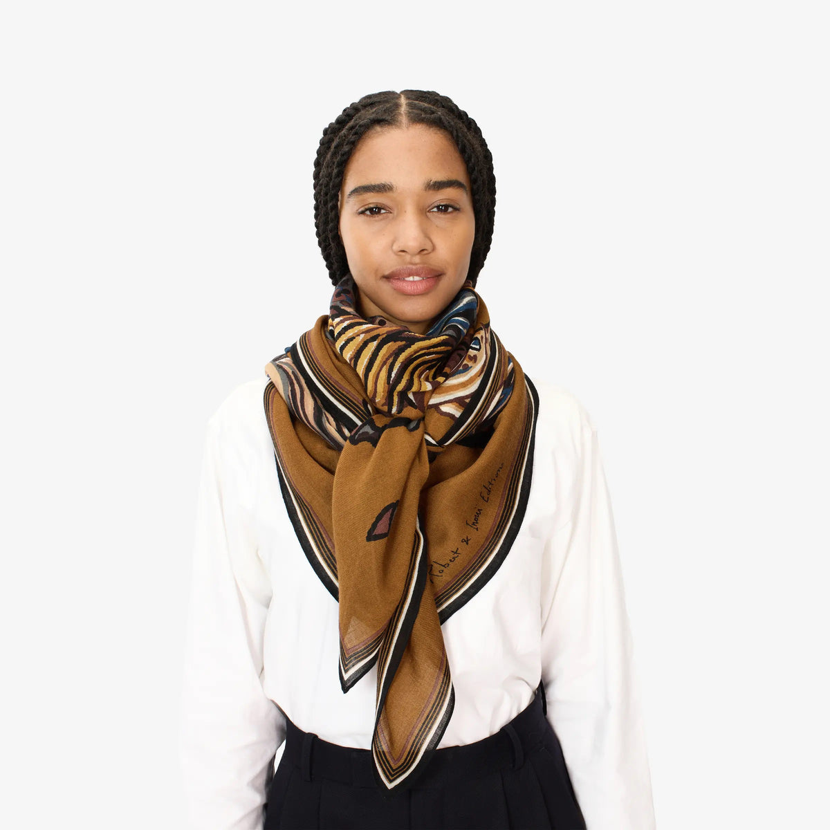 A young woman with braided hair stands against a plain white background, wearing a white shirt, black pants, and the INOUI EDITIONS - WESTERN SQUARE SCARF by INOUI, stylishly tied around her neck.