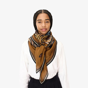 A young woman with braided hair stands against a plain white background, wearing a white shirt, black pants, and the INOUI EDITIONS - WESTERN SQUARE SCARF by INOUI, stylishly tied around her neck.