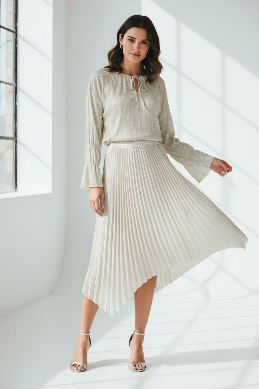 A woman with straight brown hair stands against a pale blue background, wearing the SEVENTY VENEZIA Lurex Jersey Blouse by SEVENTY with a gathered neckline and long sleeves, paired with wide-leg, high-waisted cream pants. She looks down in a relaxed pose.