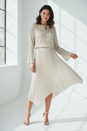 A woman with straight brown hair stands against a pale blue background, wearing the SEVENTY VENEZIA Lurex Jersey Blouse by SEVENTY with a gathered neckline and long sleeves, paired with wide-leg, high-waisted cream pants. She looks down in a relaxed pose.