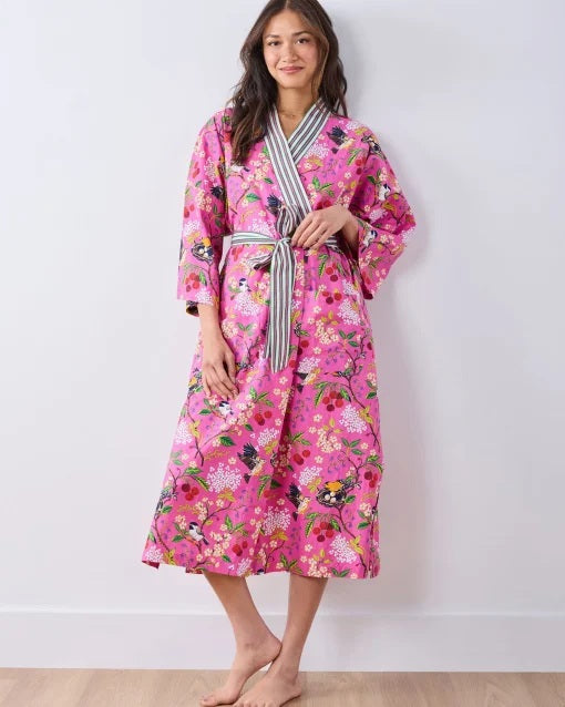 A person wearing a PRINTFRESH - BIRD & BLOSSOM LONG ROBE IN FUSCHIA, a long organic cotton robe with a bird and blossom pattern and green and white striped trim.