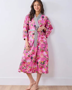 A person wearing a PRINTFRESH - BIRD & BLOSSOM LONG ROBE IN FUSCHIA, a long organic cotton robe with a bird and blossom pattern and green and white striped trim.