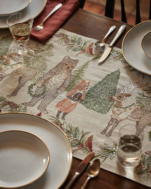 A CHRISTMAS CHEER EMBROIDERED LINEN TABLE RUNNER featuring woodland animals in winter attire and Christmas trees, draped over a wooden dining table set with plates, glassware, and candlesticks.