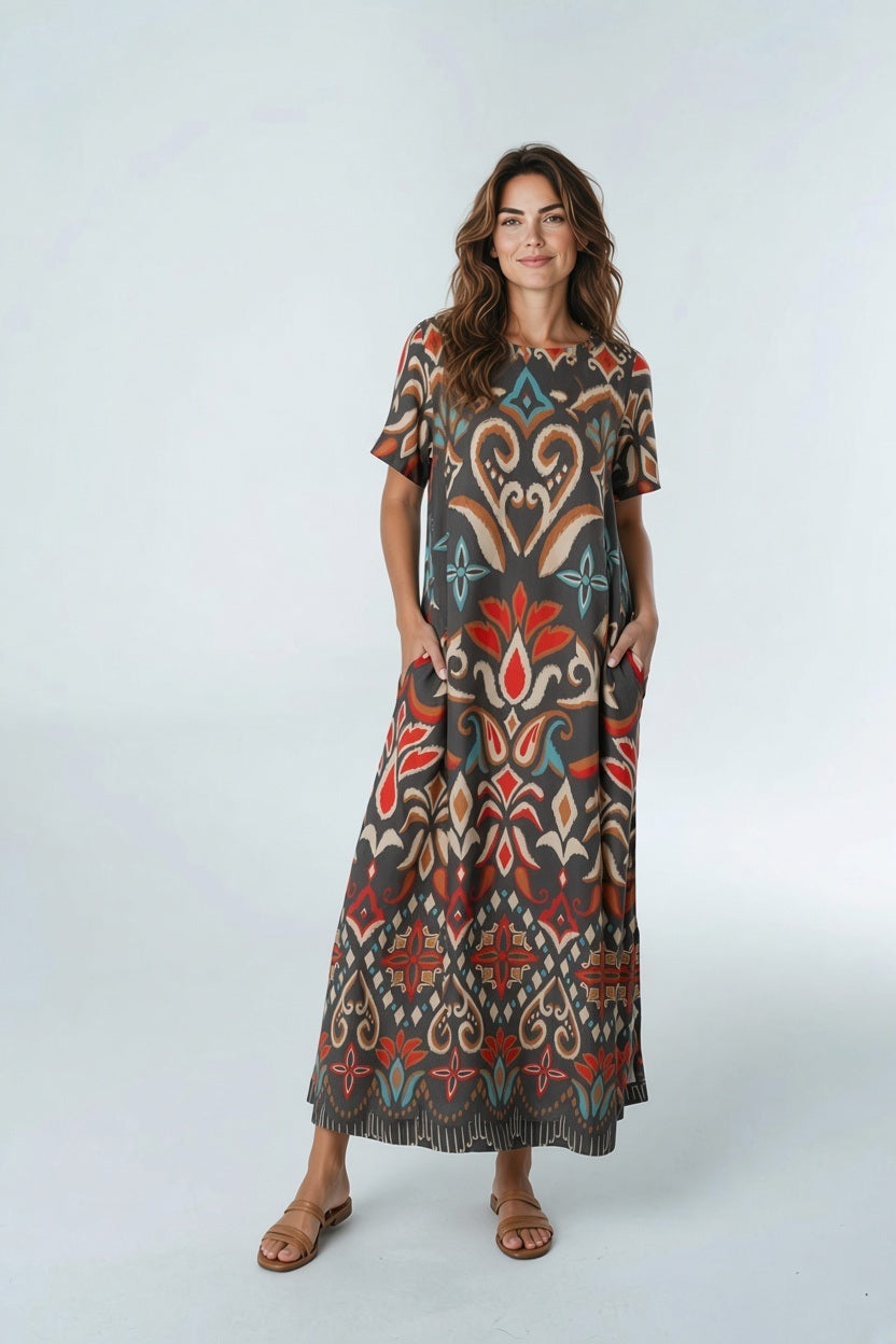 A woman wears a relaxed BRITON COURT - DIANA MAXI DRESS IN SPICED IKAT, a floor-length, short-sleeved style featuring a dark brown background with a colorful, repeating ikat-style pattern.