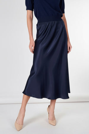 A woman with wavy brown hair wears a black short-sleeve V-neck top, black heels, and the ROSE & CROWN ROSE AND CROWN Satin Midi Skirt, standing and smiling against a plain white background.