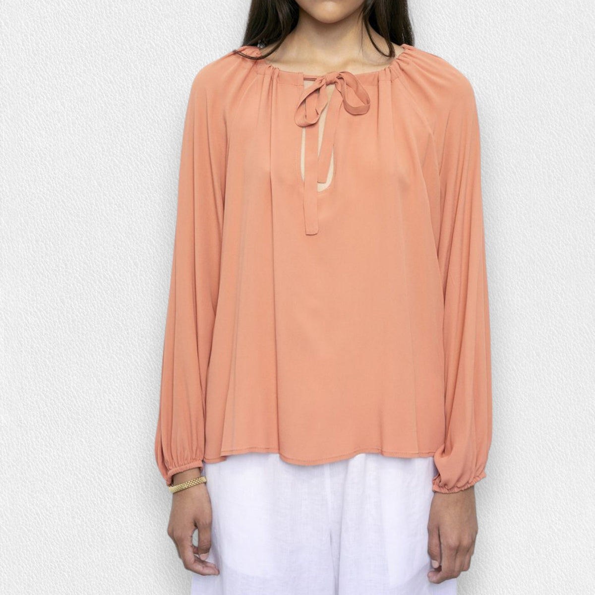A person wearing a MEIMEIJ - TIE NECK BLOUSE in salmon color, paired with white pants, stands against a light gray textured background. The blouse features long sleeves and a bow at the neckline. The person's face is not visible in the image.