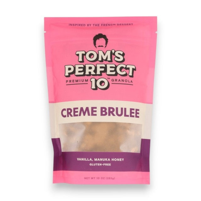 A hand pours milk over a bowl of TOM’S PERFECT 10 - CREME BRULEE GRANOLA, creating a wholesome and healthy snack. Nearby are a bag labeled “TOM’S PERFECT 10,” along with pecans, oats, manuka honey, and a small dish of crème brulee dessert on the table.