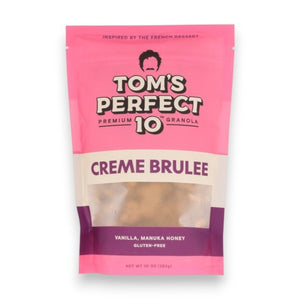 A hand pours milk over a bowl of TOM’S PERFECT 10 - CREME BRULEE GRANOLA, creating a wholesome and healthy snack. Nearby are a bag labeled “TOM’S PERFECT 10,” along with pecans, oats, manuka honey, and a small dish of crème brulee dessert on the table.