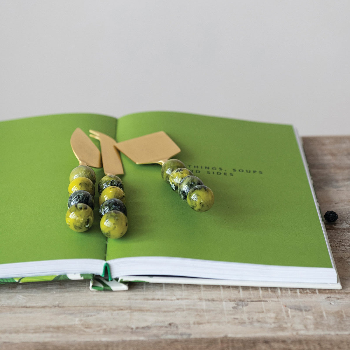 The CREATIVE COOP GOLDTONE CHEESE KNIVES WITH GREEN RESIN BALL HANDLES, SET OF 3, features three goldtone knives with marbled green handles and comes in a drawstring pouch illustrated and labeled "CHEESE KNIFE SET"—ideal for any charcuterie board.