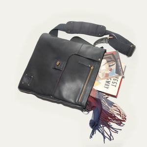 The WILL LEATHER GOODS OTTO Crossbody Bag in black full-grain leather features a front flap, gold-tone hardware, adjustable shoulder strap, and a small stitched patch on the lower right corner. Displayed against a plain white background.