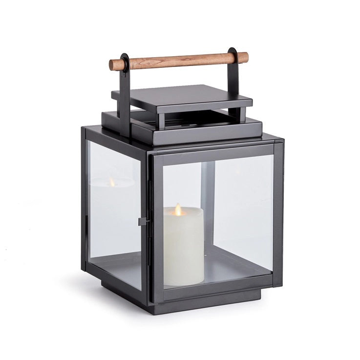 The NAPA HOME AND GARDEN - ADWIN OUTDOOR LANTERN is a black metal lantern with clear glass panels and a unique design, featuring a teak handle. It holds an unlit white candle inside, making it ideal for outdoor use with its contemporary and minimalist style.
