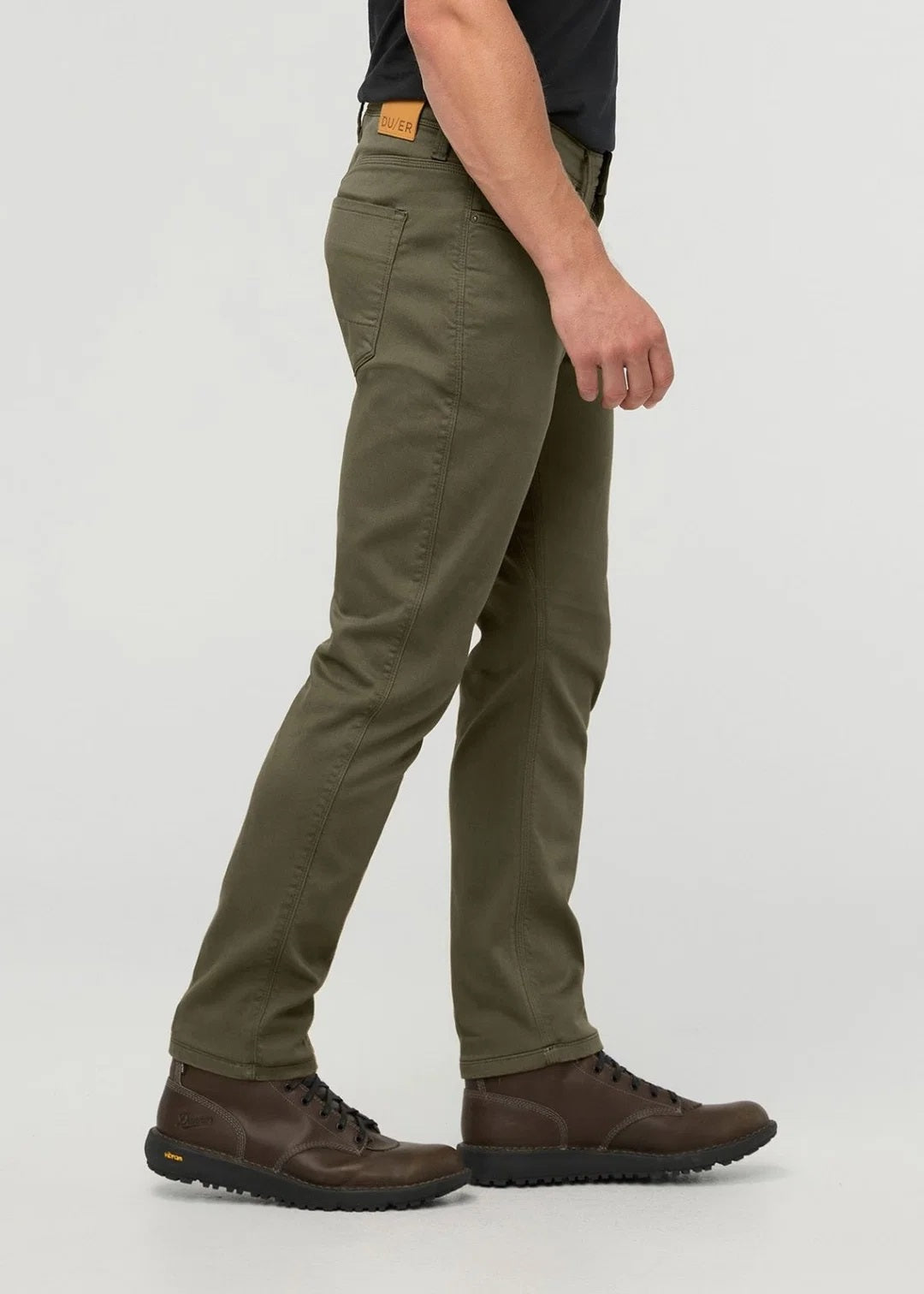 A person stands facing away from the camera against a plain background, wearing DUER - NO SWEAT ATHLETIC TAPER PANTS IN KELP paired with brown shoes.